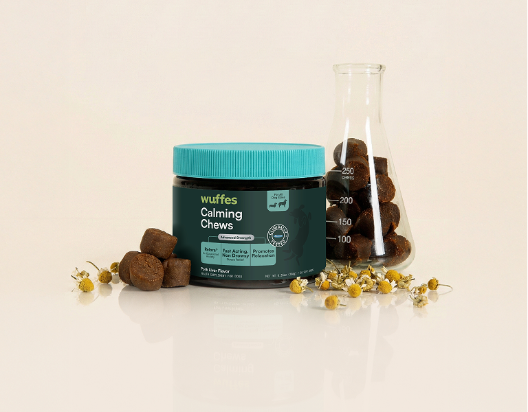 Container of calming chews with chew pieces and chamomile flowers beside a measuring flask