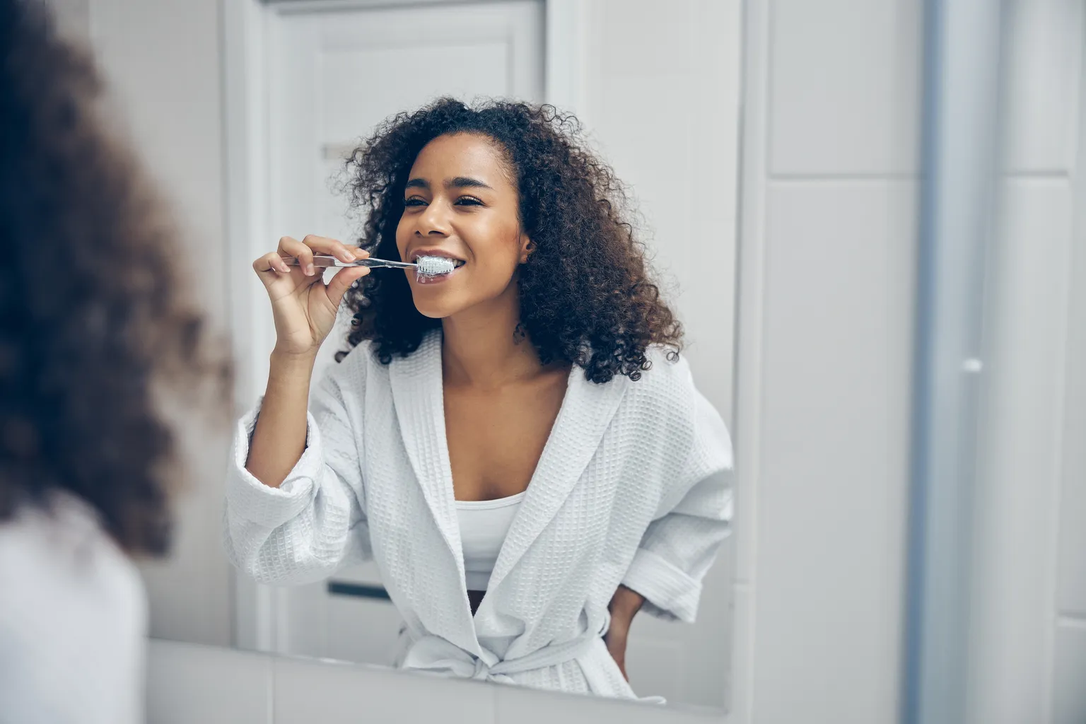 Black woman brushing her teeth in the mirror wearing white robe