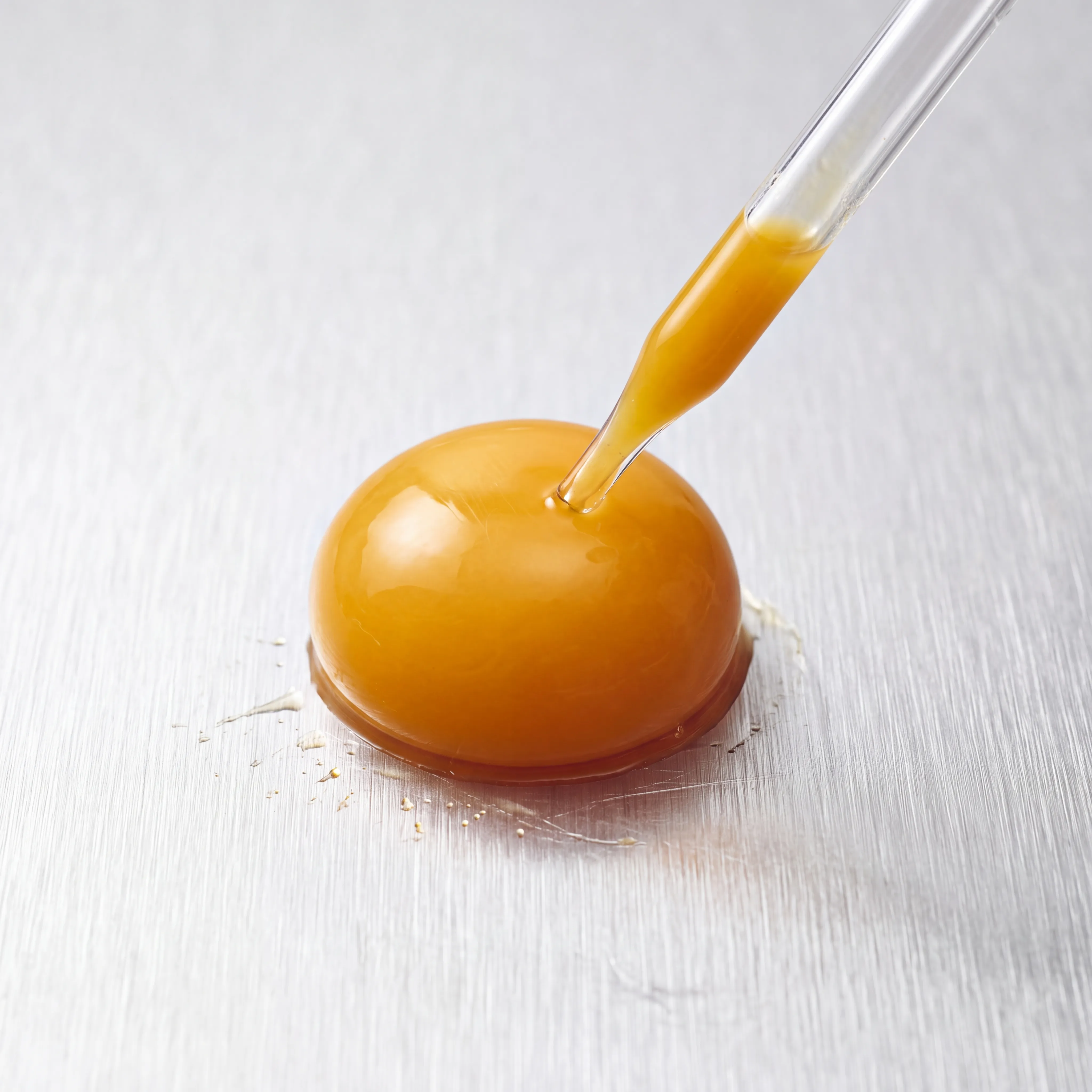 An egg yolk on brushed metal with a glass pipette dripping yolk onto it