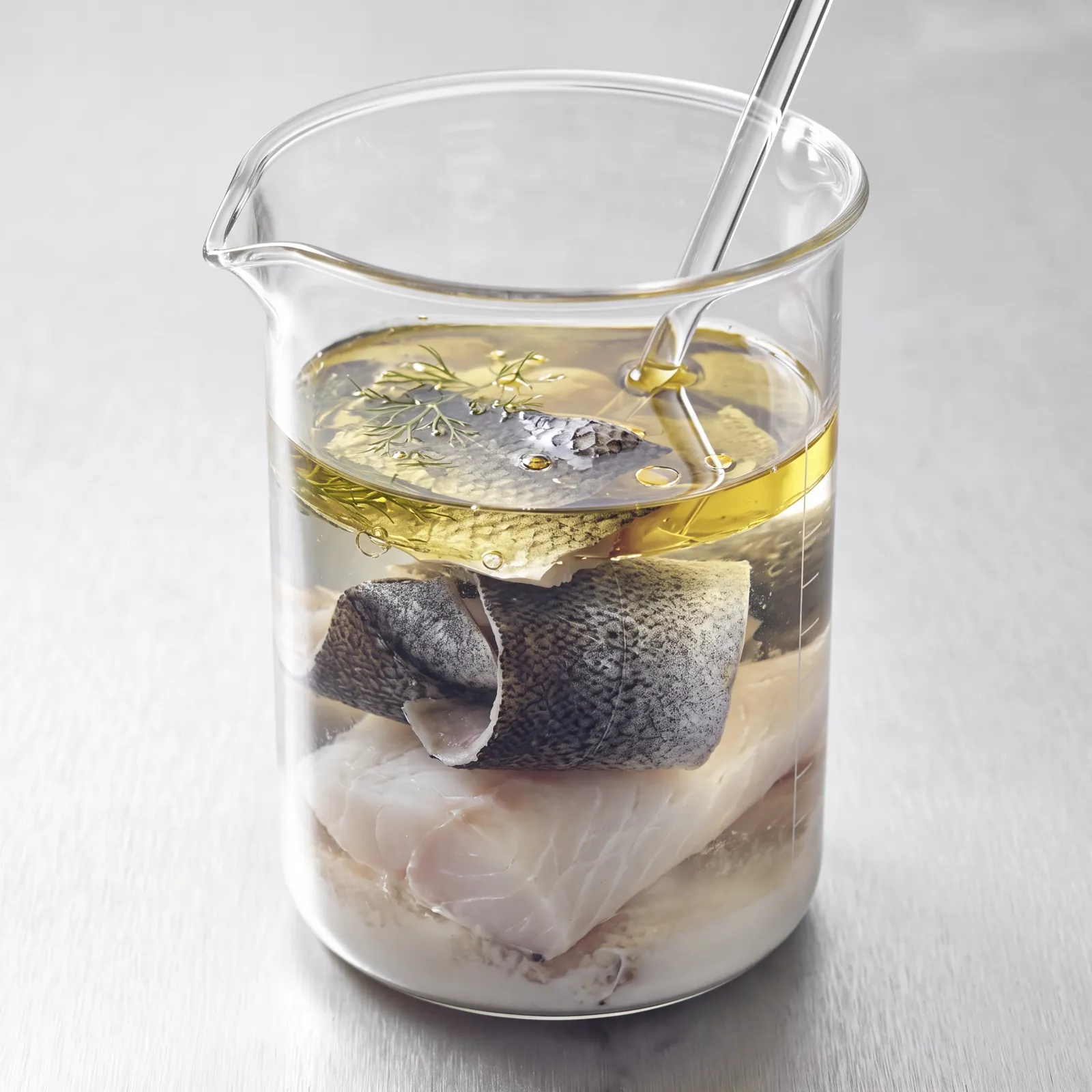 A glass laboratory beaker of golden oil holding a raw fish fillet and a rolled strip of fish skin, with a glass stirring rod resting inside