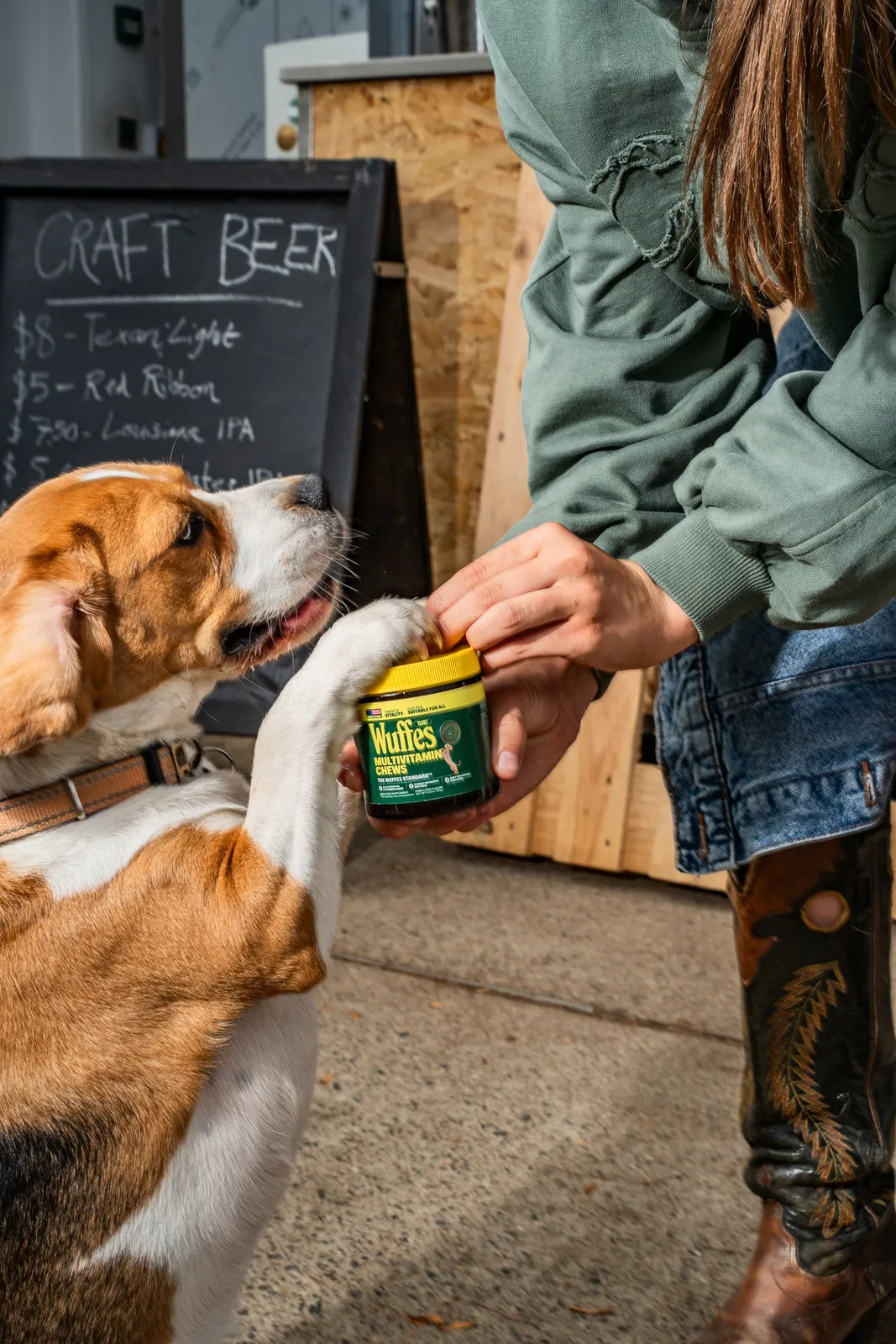 A beagle stands on its hind legs to reach a Wuffes jar held by its owner beside a chalkboard sign