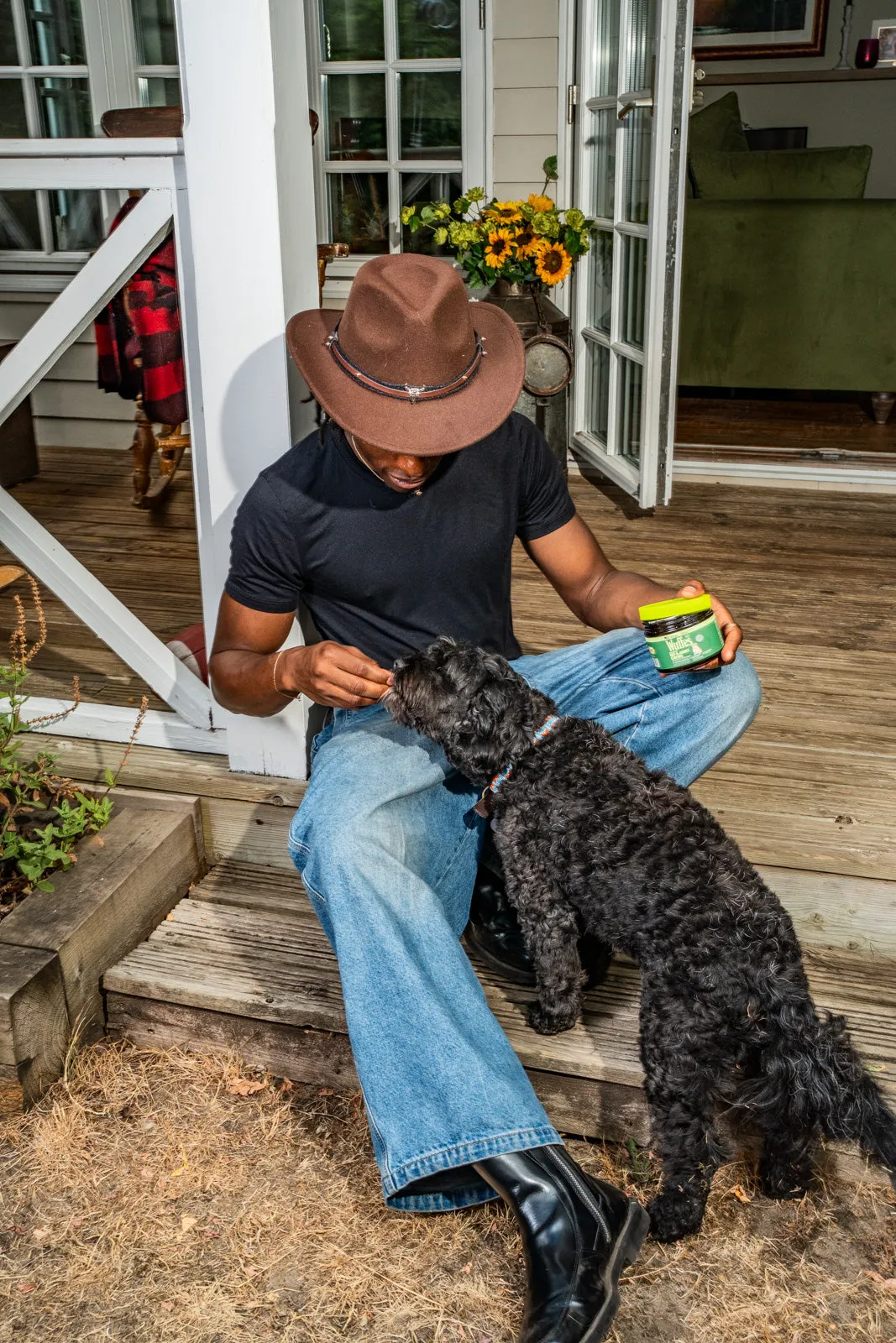 A man in a brown hat sits on wooden porch steps feeding a chew to a curly-coated black dog, a jar of Wuffes chews in his other hand