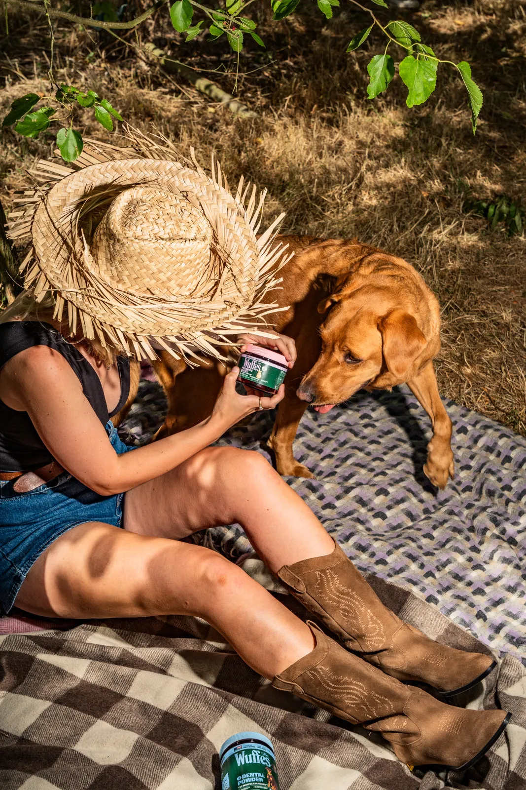 A woman in a straw hat and cowboy boots sits on a checked blanket holding a Wuffes jar beside a golden retriever