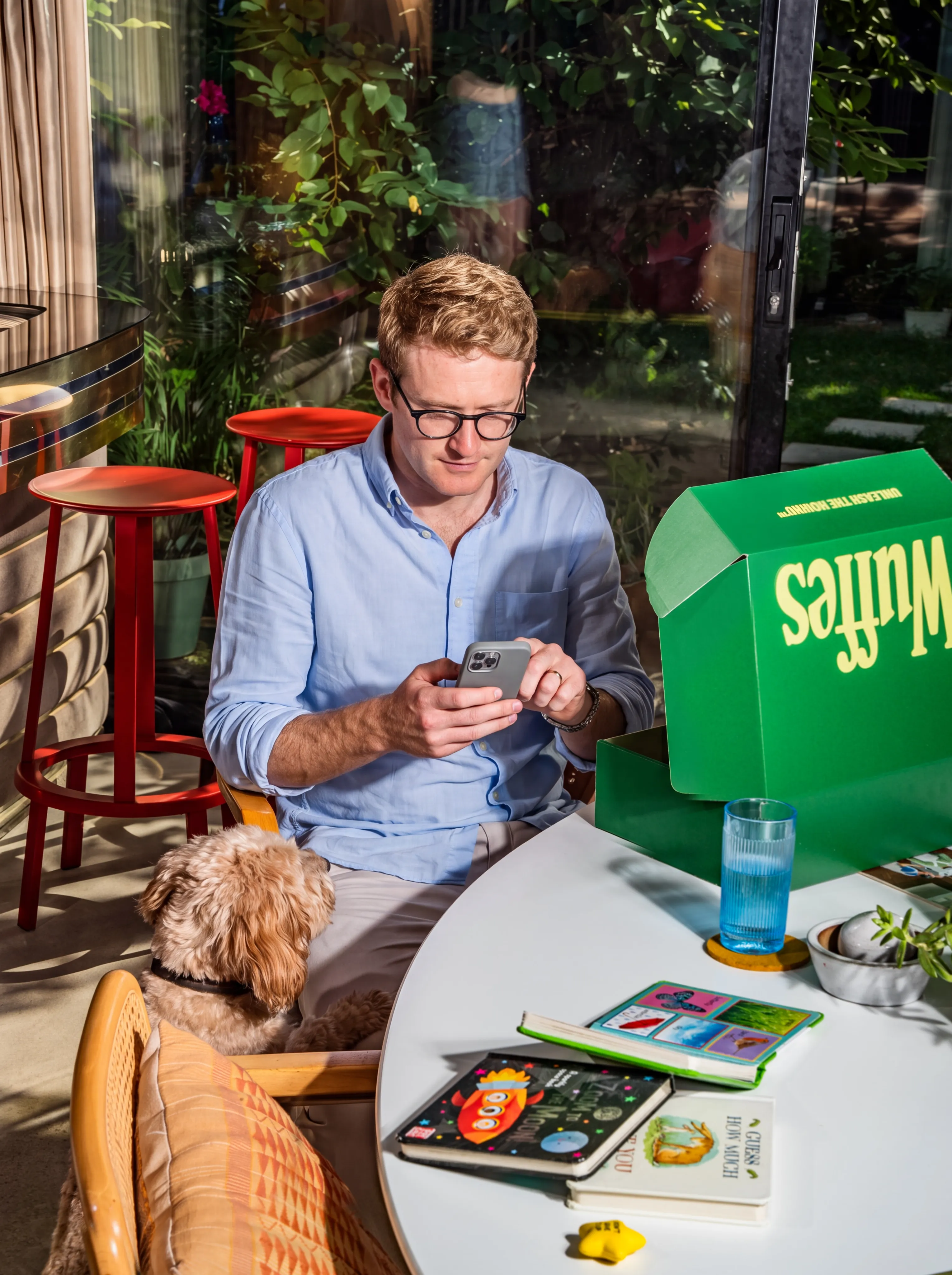 A man in glasses and a blue shirt checking his phone at a garden table beside an open green Wuffes box, with his tan cockapoo resting against his chair