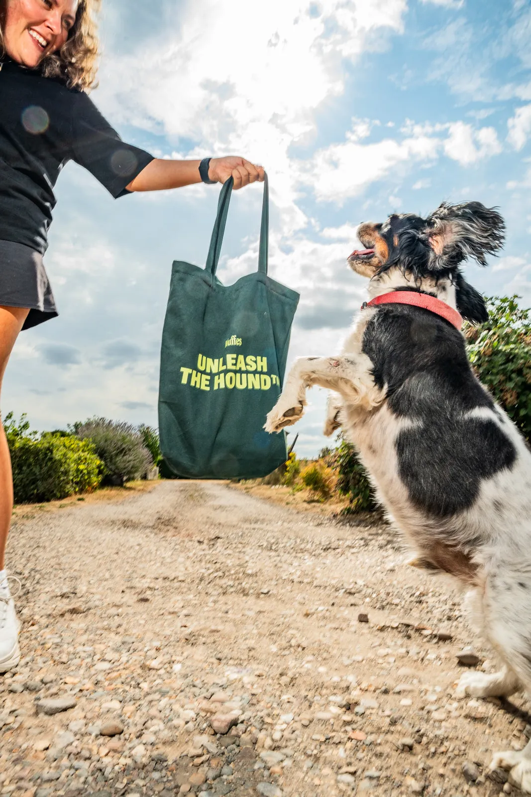 A black-and-white spaniel jumps up at a green Wuffes "Unleash the Hound" tote bag held by its owner on a dirt track