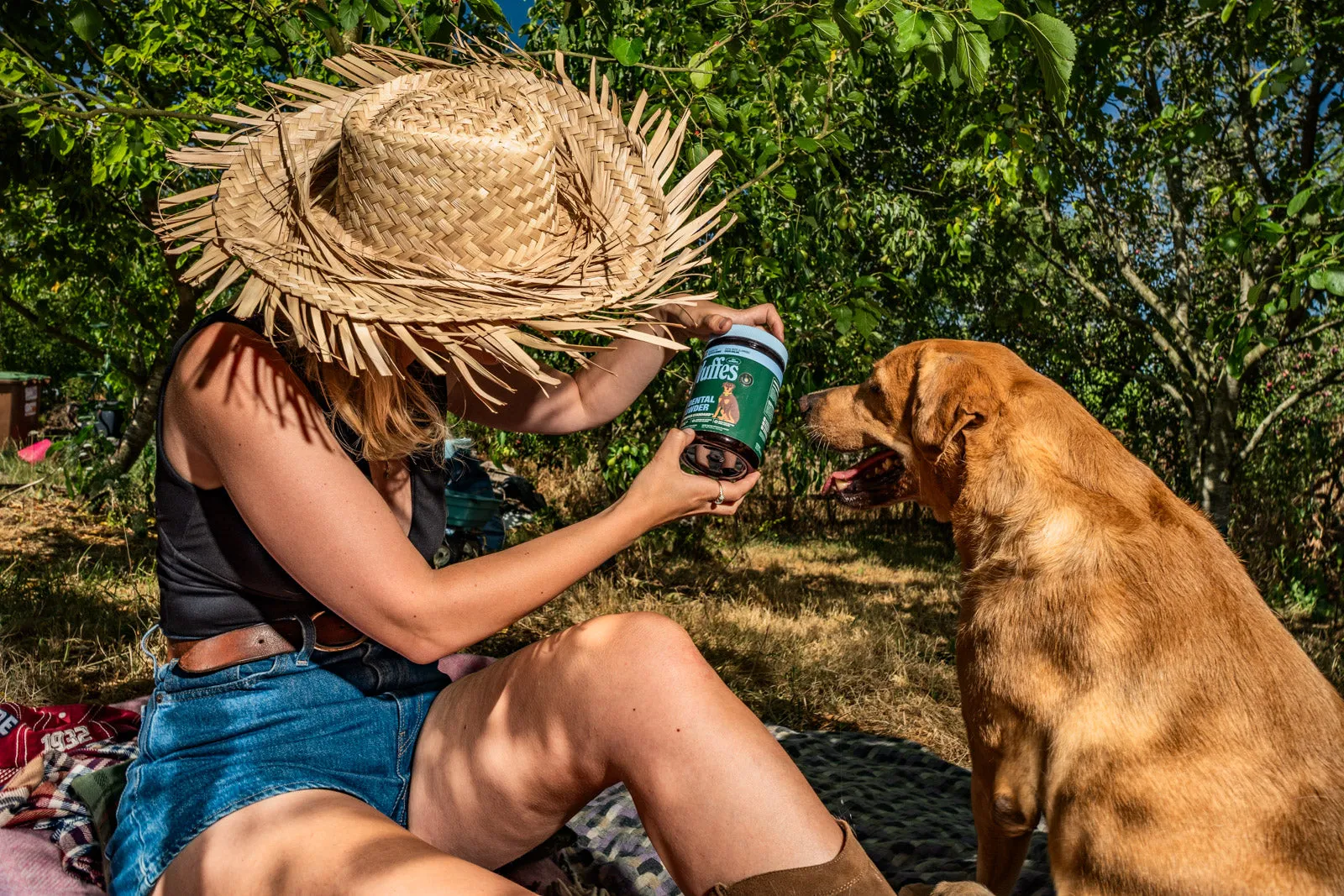 A tan short-coated dog sniffs a Wuffes jar held out by a woman in a straw hat sitting in a garden