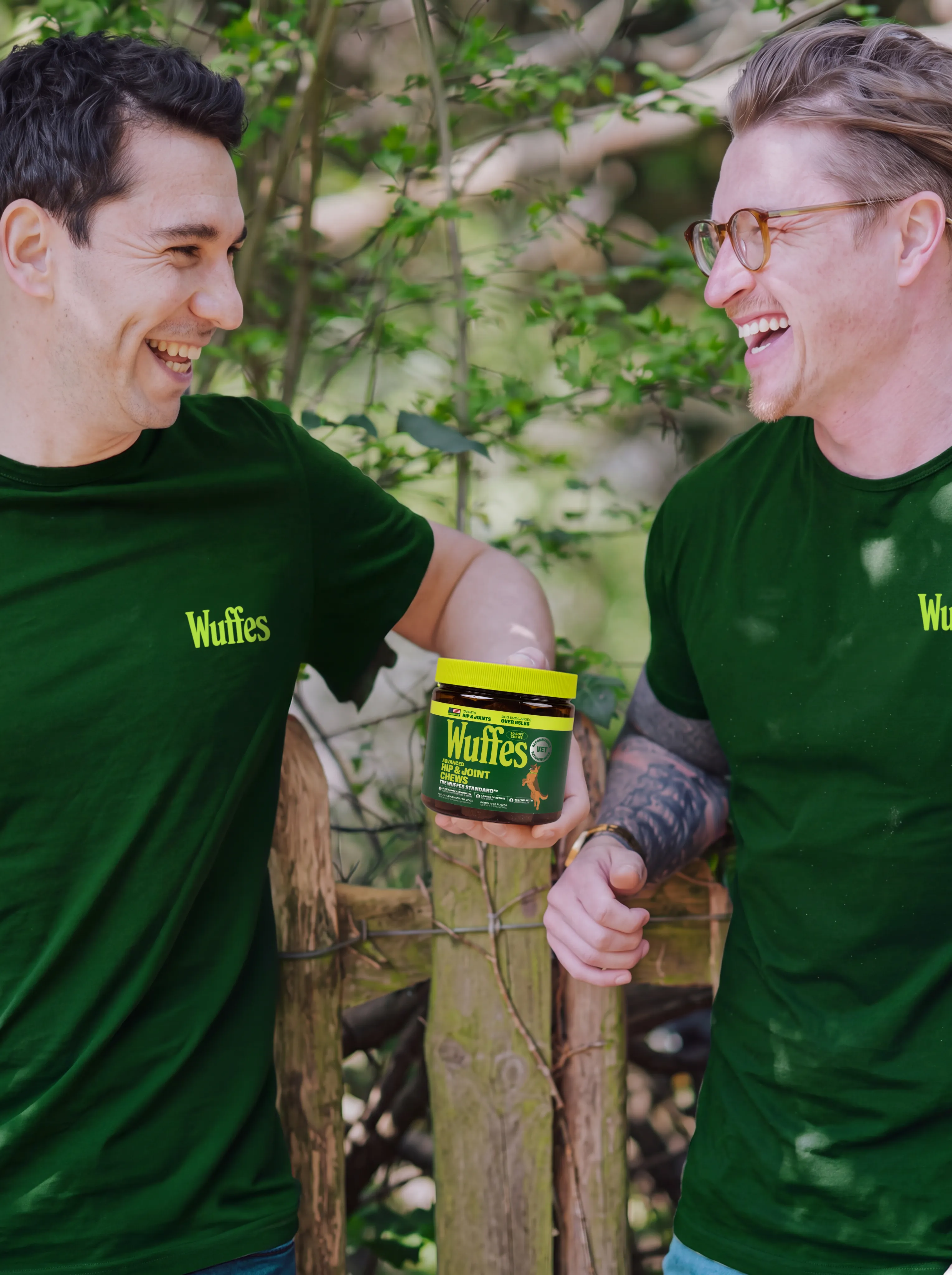 Wuffes founders Sam and Josh laughing together in green Wuffes T-shirts, one holding up a jar of Wuffes chews, beside a wooden fence under leafy trees.