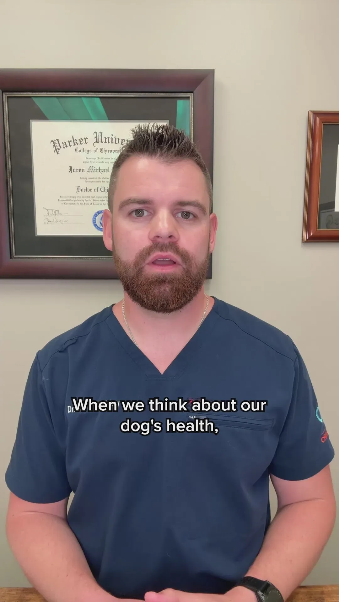 A man in navy scrubs in front of a framed diploma, talking to camera about dog health