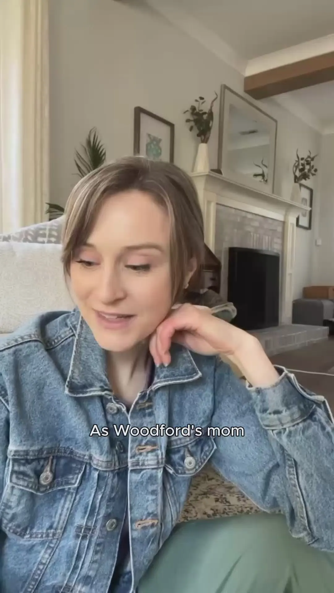A young woman in a denim jacket sitting in her living room, chin resting on her hand as she speaks to camera