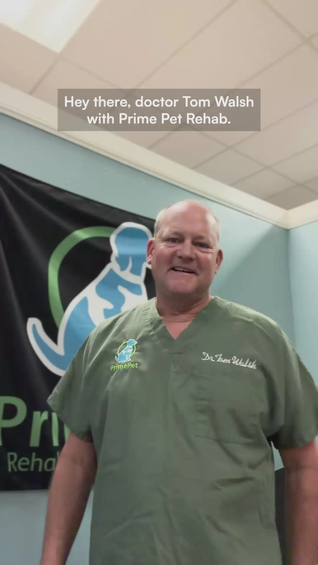 Dr. Tom Walsh of Prime Pet Rehab, in green scrubs in front of the clinic banner, introducing himself to camera