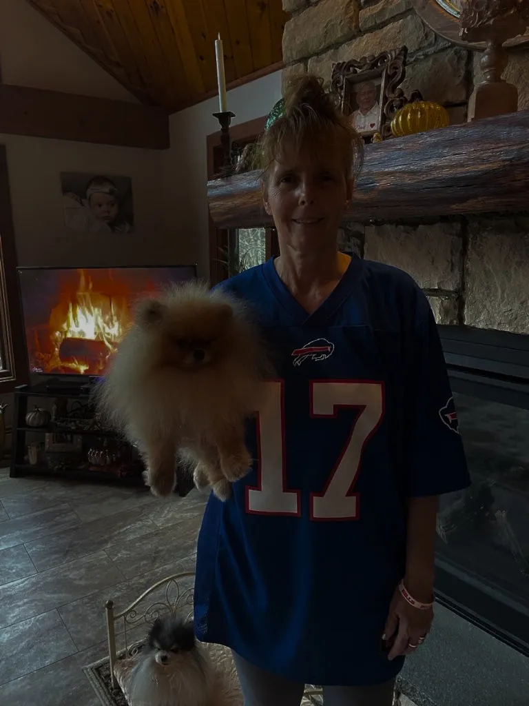 A woman in a blue number 17 football jersey holds up a fluffy orange Pomeranian in a living room with a lit fireplace
