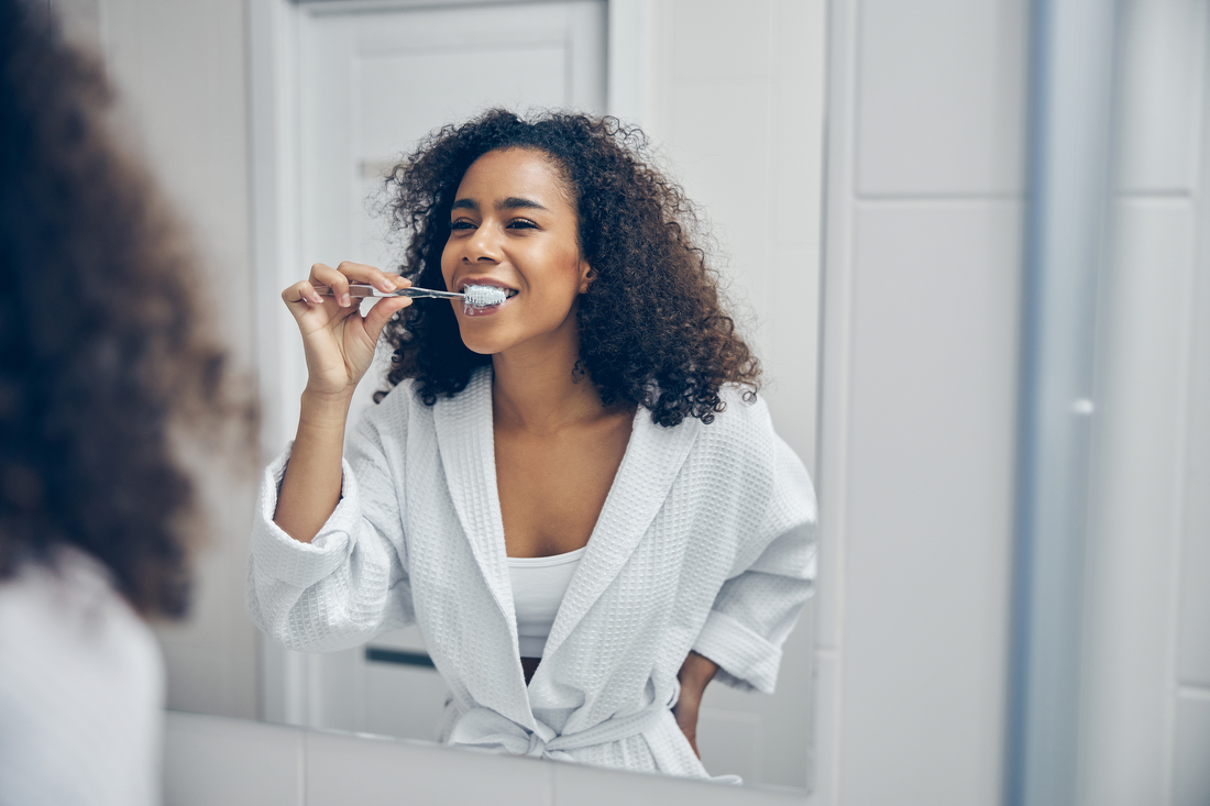 Black woman brushing her teeth in the mirror wearing white robe